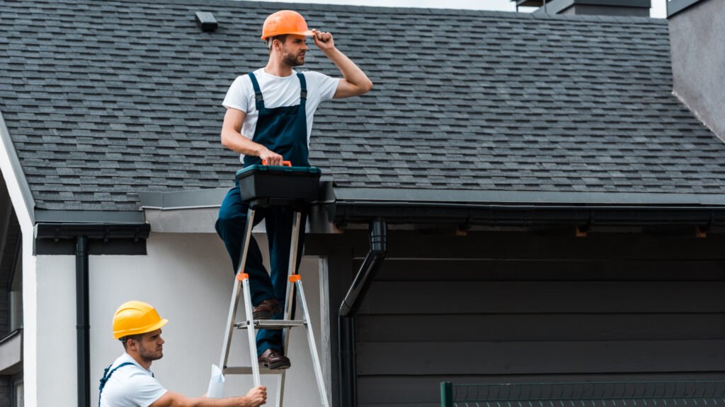 handsome repairman sitting on roof and holding toolbox near coworker in helmet